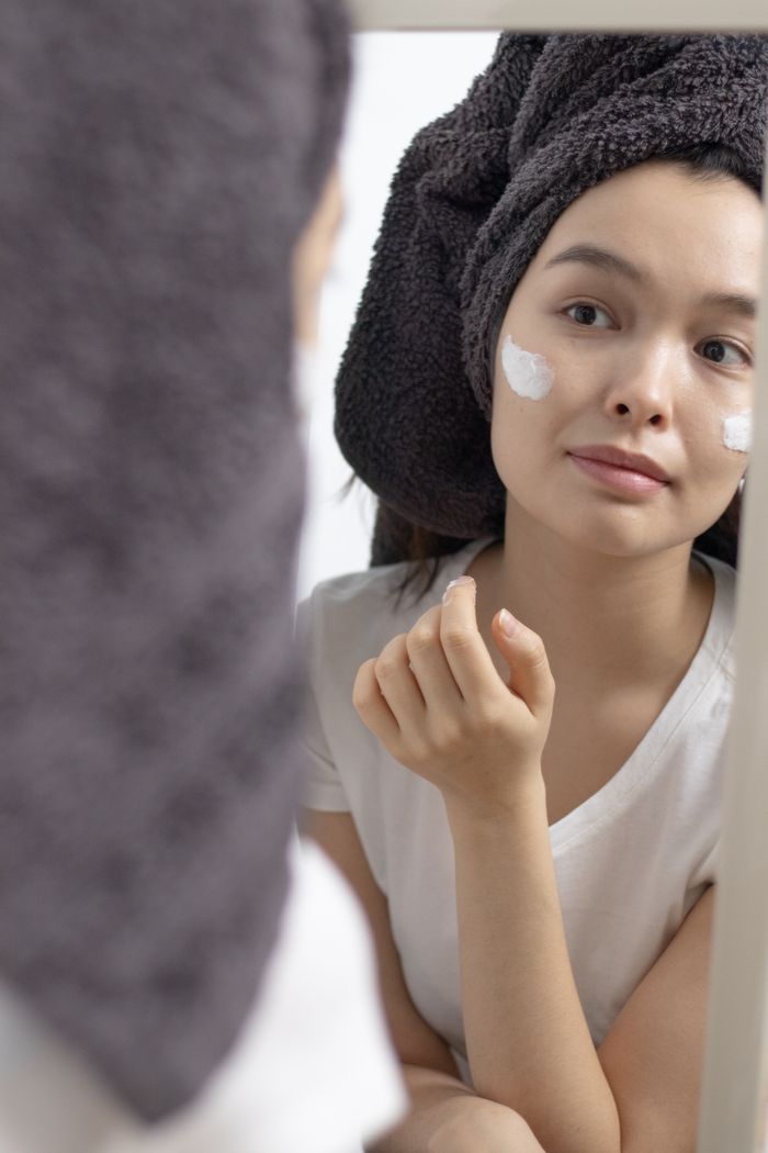 services-01 Woman in towel applying face cream, looking at her reflection. Skincare routine in the bathroom.
